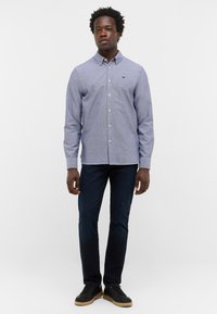 Hemd mit Knöpfen aus hellblauem Stoff mit dezentem Muster, langen Ärmeln, Schulterpasse und kleinem Logo auf der Brust. Getragen mit dunkler Hose.