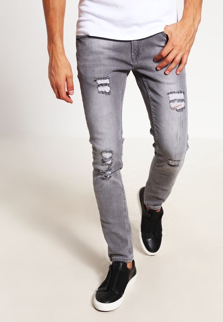 Homme marchant portant un jean gris déchiré, des chaussures slip-on noires avec des semelles blanches et une chemise blanche, montré de la taille aux pieds.