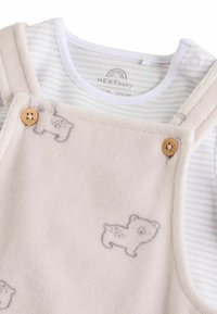Fleece overallit vaaleassa beessissä, jossa on karhukuosi, puunapit ja raidallinen valko-harmaa pitkähihainen paita alla.