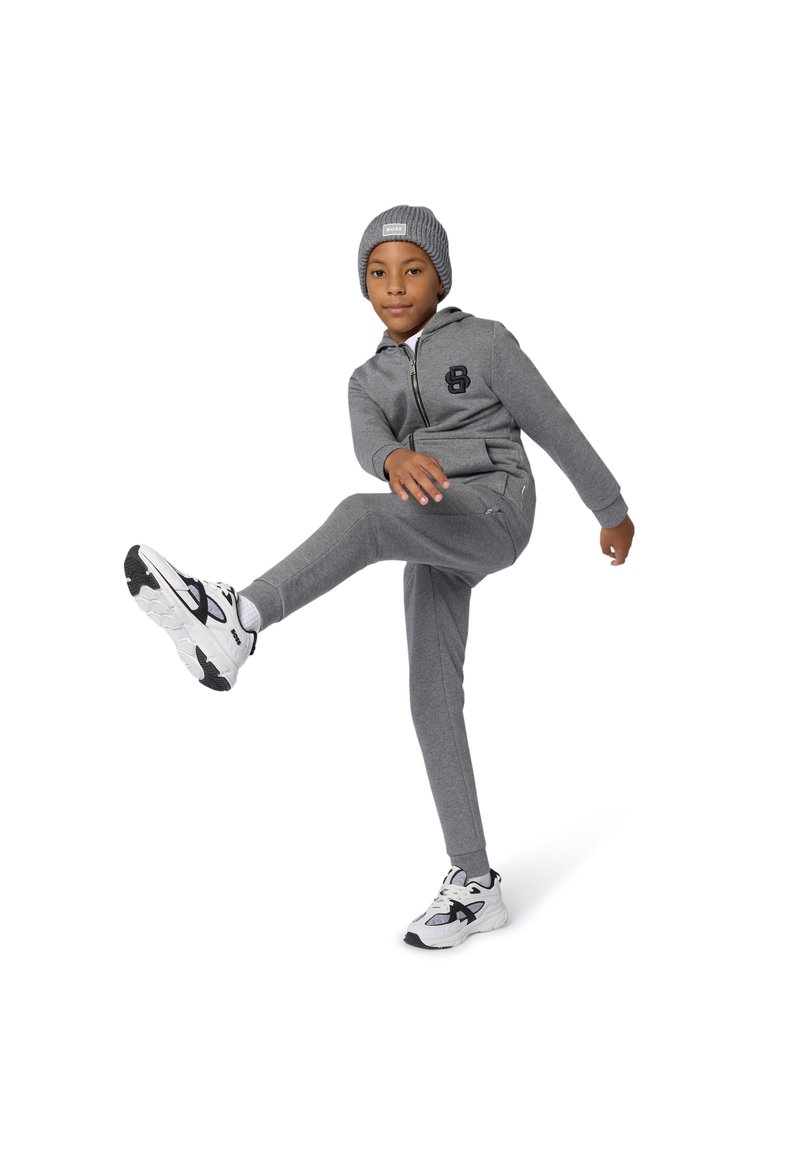 Siva jopa s kapuco z zadrgo in ujemajoče trenirka hlače s črnim logotipom na prsih, kombinirano z belimi in črnimi športnimi čevlji. Teksturirana pletena kapa.