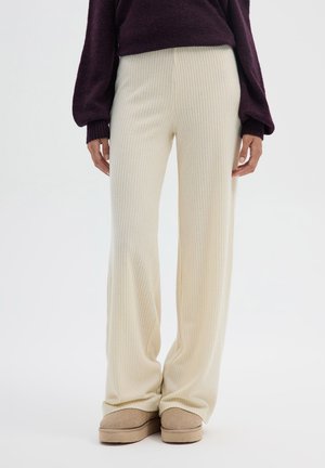 Persona che indossa pantaloni larghi a coste color crema, un maglione a maniche lunghe viola scuro e scarpe beige slip-on, in piedi davanti a uno sfondo neutro.