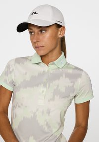 Jasnozielona koszulka polo z abstrakcyjnym wzorem, krótkimi rękawami i zapięciem na guziki. Biała czapka z czarnym logo i daszkiem.