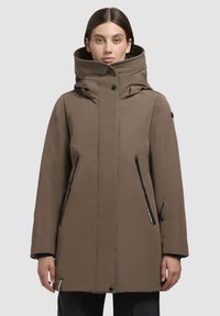 Femme portant un manteau d'hiver marron jusqu'aux genoux avec un col haut et des poches zippées, se tenant devant un fond clair.