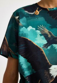 T-shirt med kort ärm i mörk teal med detaljerade örn grafik och stiliserade moln, som visar ett naturligt tema.