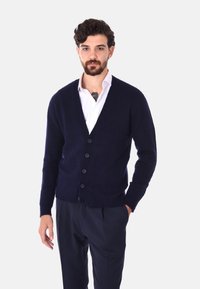 Cardigan blu navy realizzato in morbida filato con scollatura a V, polsini a coste, sei bottoni sul davanti e vestibilità comoda. Indossato sopra una camicia bianca.
