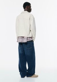 Jachetă din denim alb supradimensionată, purtată peste o cămașă cu carouri, completată cu blugi largi de culoare albastră închis. Design simplu, cusături minime și detalii subtile din metal.
