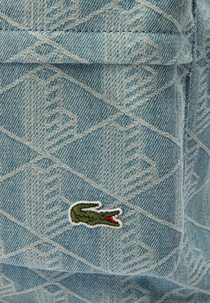 Tissu en denim avec un motif en losanges et un logo crocodile vert brodé, bouche rouge ouverte, sur une poche.