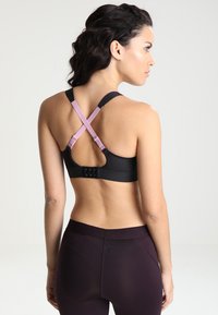 Svart sports-bh med justerbare rosa stropper og en krok-lukking bak. Har en glatt tekstur og krysset stroppdesign.