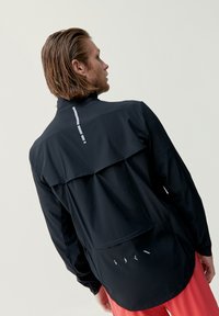 Svart idrottsjacka med hög krage, med en lagerpålagd baksidedesign, reflexdetaljer och en lätt textur.