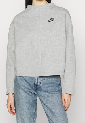 Osoba ubrana w jasnoszarą bluzę Nike z czarnym logo i niebieskie dżinsy, widziana od ramion do bioder.