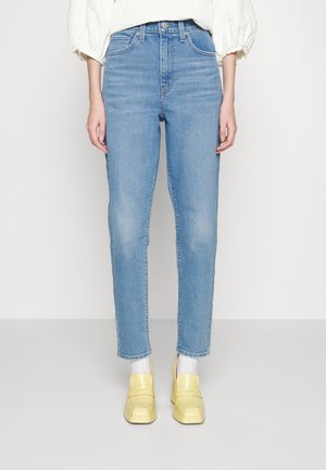 Personne portant un jean slim bleu clair, des chaussettes blanches, des mocassins jaunes et un chemisier blanc à manches longues, debout devant un fond uni.
