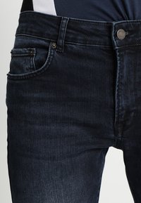 Vaqueros de mezclilla azul oscuro en la cintura y la parte superior del muslo con botón visible, presillas para cinturón, bolsillos frontales y bolsillo para monedas, combinados con una parte superior en azul marino y blanco.