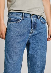 Jeans de mezclilla azul claro con un corte relajado, que cuentan con estilo de cinco bolsillos, trabillas para cinturón y cierre de botón metálico. Presenta un sutil desgaste en la tela.