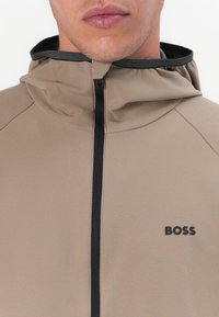 Tan hoodie met een volledige rits, met een zwarte rits en logo-detail. De stof lijkt glad met een licht matte afwerking.