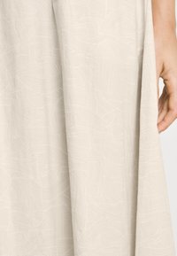 Ljusbeige tyg med abstrakta, vågiga linjemönster. Slät textur med draperade veck och en hand som är placerad nära kanten.