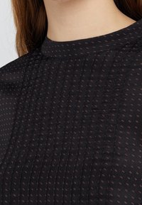 Svart blus med hög ringning, med en struktur av vertikala veck och små, kontrasterande mörkröda prickar i hela tyget.