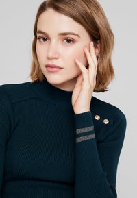 Petrolkleurig geribbelde trui met een hoge hals, voorzien van gouden knoopaccenten op de schouder en metalen strepen op de manchetten. Zachte textuur.