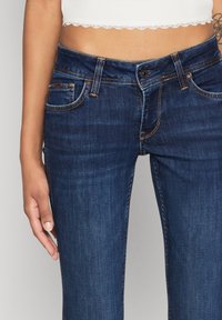 Ciemnoniebieskie dżinsy o slim fit, z miedzianym przeszyciem, czterema kieszeniami oraz zapięciem na guzik w pasie.