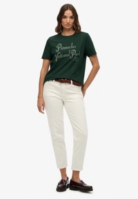 Mørkegrøn bomulds T-shirt med "Pinnacles National Park" tryk, parret med hvide jeans og et brunt vævet bælte; modellen bærer sorte loafers.