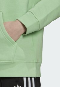 Ljusgrön sweatshirt med en kenguruficka och ribbad mudd; slät textur och avslappnad passform. Svarta byxor med vita ränder synliga.