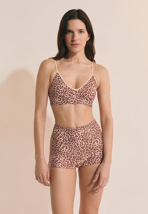 Frau trägt einen passenden Leopardenmuster-Bralette und High-Waist-Shorts, steht vor einem schlichten beigen Hintergrund.