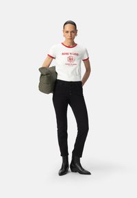 Camiseta blanca de manga corta con ribetes rojos, que presenta un gráfico de una rosa y texto. Se lleva con jeans de tiro alto negros y botas negras hasta el tobillo. Sosteniendo una chaqueta verde.