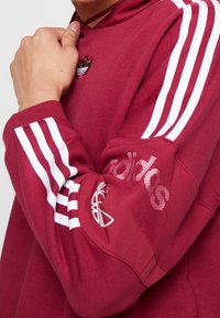 Vinröd huvtröja med vita ränder på ärmarna, ribbade muddar och texturerade detaljer som har en logotyp och märket "adidas" på armen.