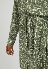 Olivgrön klänning med ett vitt ojämnt prickmönster, långa ärmar, knappfront och en tygbälte knuten i midjan.