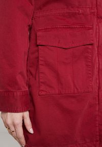 Giacca rossa con una grande tasca quadrata con pattina, realizzata in tessuto testurizzato con una leggera lucentezza. Cuciture visibili lungo le cuciture.