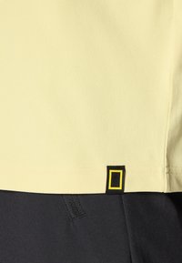 Yellow short-sleeve shirt made of soft fabric, featuring a black label with a yellow square logo at the hem. Black pants with textured detail.