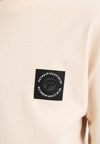 Felpa beige chiaro con una texture liscia, caratterizzata da un quadrato nero con l'etichetta "Petrol Industries" in un testo circolare bianco.