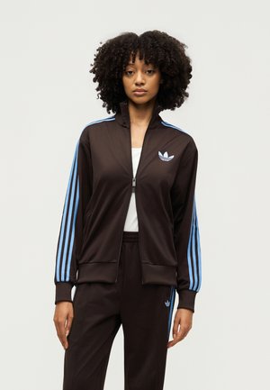 Frau mit lockigem Haar, die einen schwarzen Adidas-Trainingsanzug mit hellblauen Streifen und Logo trägt, steht vor einem schlichten hellen Hintergrund.