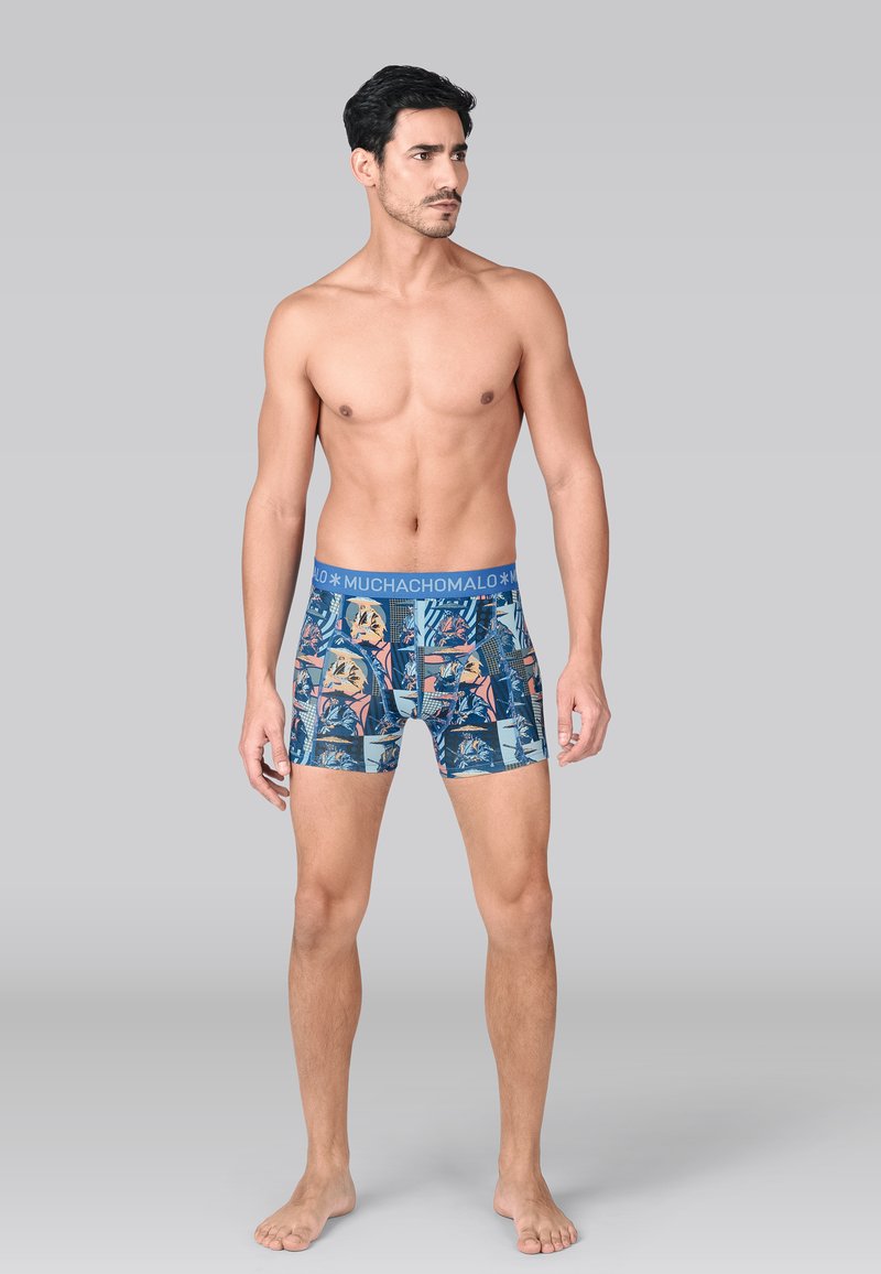Shirtless man wearing blue patterned boxer briefs with tropical prints and "MUCHACHOMALO" waistband, standing barefoot on gray background.