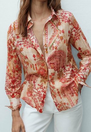 Vrouw die een blouse met lange mouwen met rode en oranje bloemenpatronen en een witte broek draagt, staand tegen een effen lichte achtergrond.