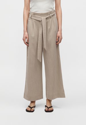 Femme portant un pantalon beige à jambes larges avec une ceinture en tissu nouée et des sandales noires à bout ouvert, debout devant un fond uni.