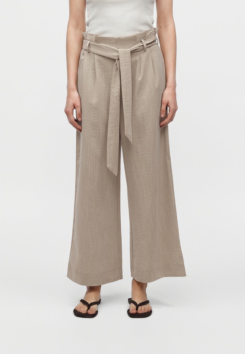 Mujer con pantalones beige de pierna ancha con un cinturón de tela atado y sandalias negras abiertas, de pie frente a un fondo liso.