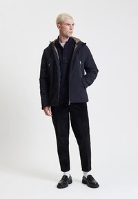 Giacca nera isolante con cappuccio beige, tasche con zip e finitura liscia, abbinata a pantaloni in velluto a coste neri e mocassini neri.