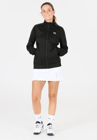 Svart zip-up jacka med hög krage, FZ-logotyp och mönstrade detaljer. Kombineras med en vit sportkjol och vita träningssko.