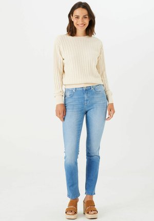 Lichtcrème geribbelde gebreide trui met lange mouwen, gecombineerd met lichtblauwe slim-fit jeans en bruine platform sandalen. Effen achtergrond.