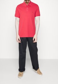 Polo shirt in cotone rosso con colletto a punta, patta con due bottoni e dettaglio del logo, abbinato a pantaloni neri e sneakers beige.