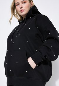 Sudadera negra hecha de tela suave, con un diseño de gran tamaño, un bolsillo delantero y adornada con pequeñas perlas blancas en toda su superficie.