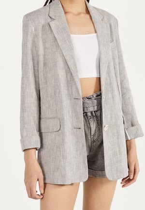 Frau trägt einen hellgrauen Blazer über einem weißen Cropped-Top und hoch taillierte graue Jeansshorts mit aufgerollten Säumen.