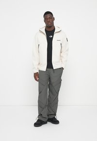Lys beige zip-jakke med to forlommer, sort t-shirt under, mørkegrå cargo-bukser med flere lommer, sorte sko.