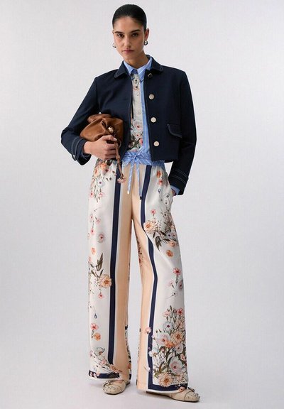 Femme portant une veste courte bleu marine, une chemise bleue à col, un pantalon large à imprimé floral, tenant un sac en cuir marron sur un fond uni.