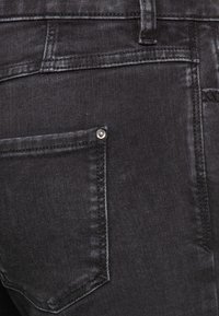 Jeans de mezclilla negra con una textura suave y un brillo sutil, que cuentan con un bolsillo trasero con un remache metálico y costuras en contraste.