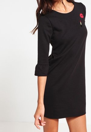 Femme portant une robe noire à manches trois-quarts, ornée d'une petite tache de lèvres rouges et de rouge à lèvres près de l'épaule gauche.