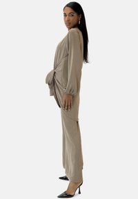 Abito beige a maniche lunghe con dettaglio annodato drappeggiato in vita e vestibilità comoda. Abbinato a décolleté nere con punta appuntita. Tessuto dalla texture liscia.