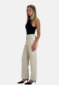 Zwart mouwloos topje, beige high-waisted broek met een gladde textuur en zijrits, gecombineerd met zwarte puntige hakken.