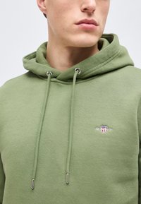 Νέος άνδρας που φοράει ανοιχτό πράσινο φούτερ με κορδόνια και ένα μικρό κεντημένο λογότυπο στο στήθος, κοιτάζει ελαφρώς πλάγια.