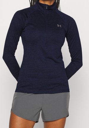 Kvinde iført en marineblå atletisk langærmet top med kvart lynlås og grå atletiske shorts, står med hænderne bag ryggen.